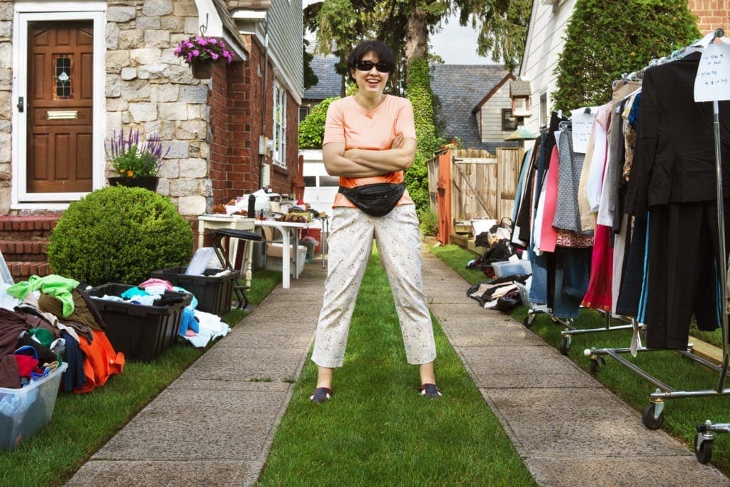 A smiling Mom standing with her arms crossed in her drive way after setting up her yard sale