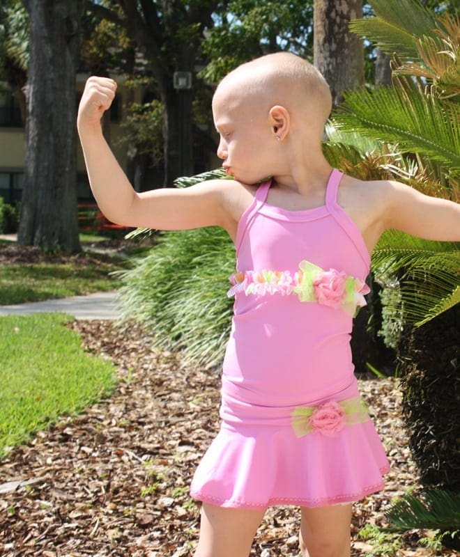 Picture of young little girl in pink ballerina outfit flexing to kiss her bicep so that she is strong and can beat cancer